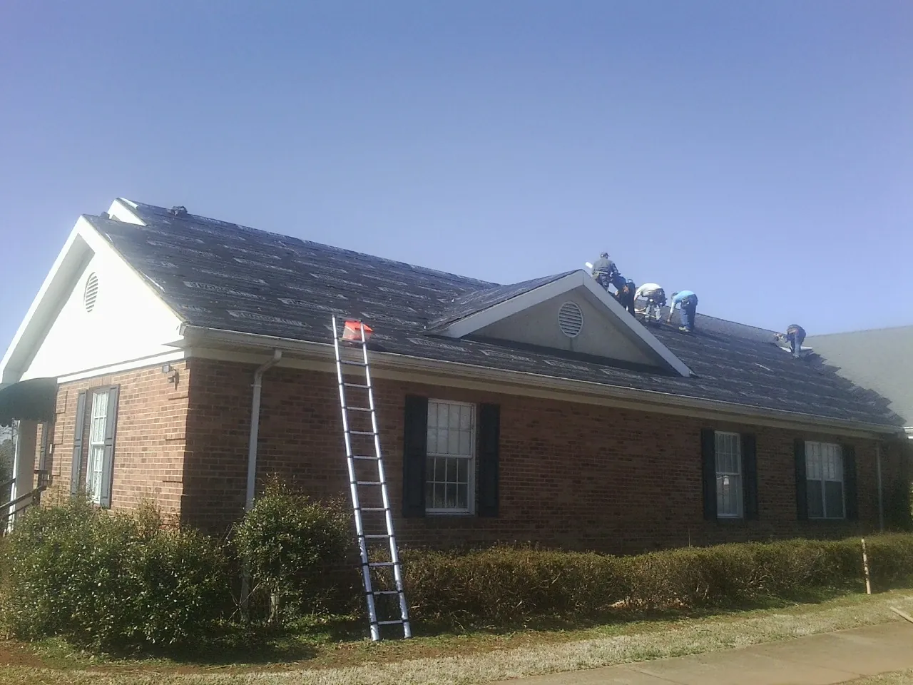 Crew tearing shingles off a brick ranch roof with a ladder set — Emergency Roof Repair work by our crew in Ickesburg
