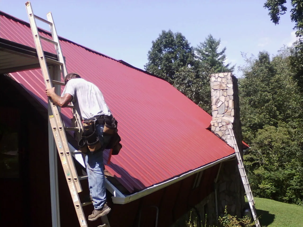 Roofer on a ladder beside a red metal roof and stone chimney — Soffit & Fascia Repair by Roofing Brothers in Ickesburg